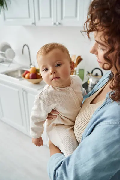 Femme heureuse tenant mignonne petite fille dans la barboteuse tout en se tenant dans la cuisine à la maison, mère aimante — Photo de stock