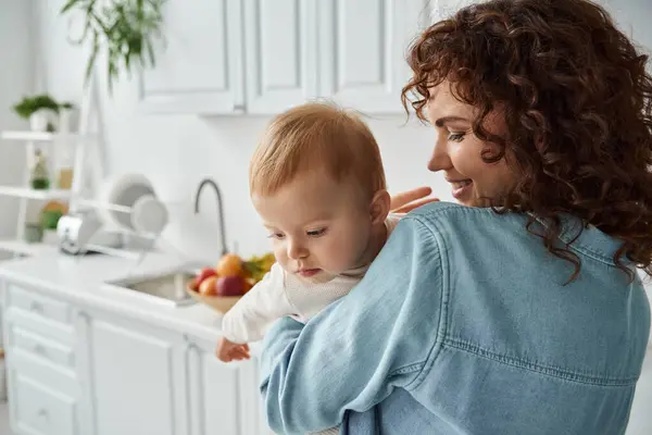 Donna allegra con i capelli ondulati che tengono la figlia del bambino in cucina a casa, felice maternità — Foto stock