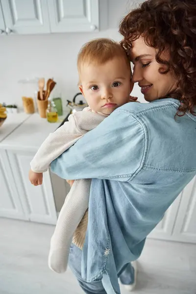 Bébé fille regardant caméra dans les mains de la mère avec les yeux fermés l'embrassant dans la cuisine à la maison — Photo de stock