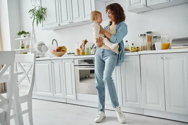 Longitud completa de la mujer feliz sosteniendo hija pequeña en la cocina moderna espaciosa con muebles blancos - foto de stock