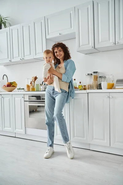 Longitud completa de la mujer sonriente con la pequeña hija mirando a la cámara en la cocina con el interior blanco - foto de stock