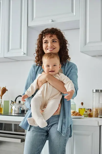 Femme heureuse avec les cheveux ondulés tenant fille tout-petit et souriant à la caméra dans la cuisine moderne à la maison — Photo de stock