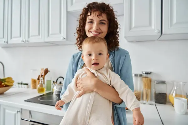 Amorosa madre con el pelo ondulado abrazando adorable hija en mamada en casa en la cocina, felicidad familiar - foto de stock