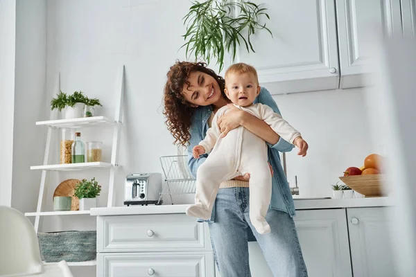 Donna allegra che tiene piccola figlia in pagliaccetto in cucina con interni contemporanei, casa accogliente — Foto stock