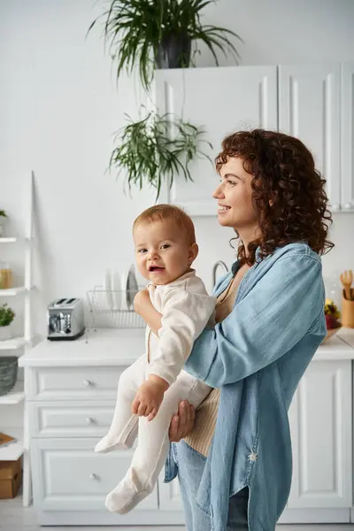 Bambino allegro in pagliaccetto che guarda la macchina fotografica in mani di madre in cucina a casa, infanzia felice — Foto stock
