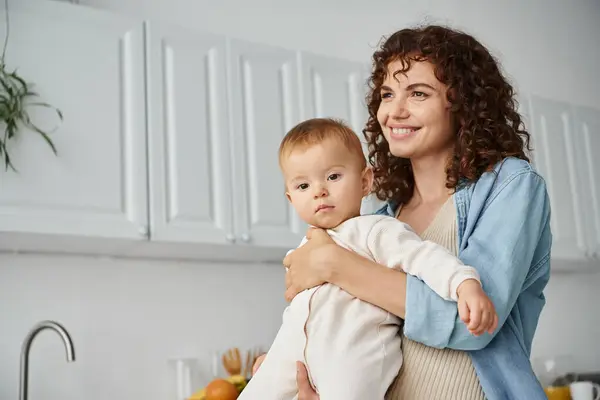 Donna con i capelli ricci e sorriso felice guardando lontano vicino figlia bambino in cucina, maternità — Foto stock