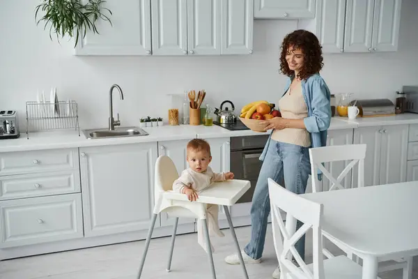 Smiling woman with bowl of fresh fruits near child in baby chair in modern kitchen, morning mealtime — Stock Photo