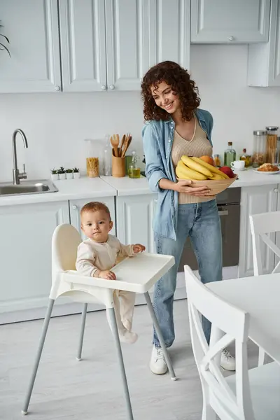 Joyful woman holding bowl of fresh fruits near kid in baby chair in contemporary kitchen, breakfast — Stock Photo