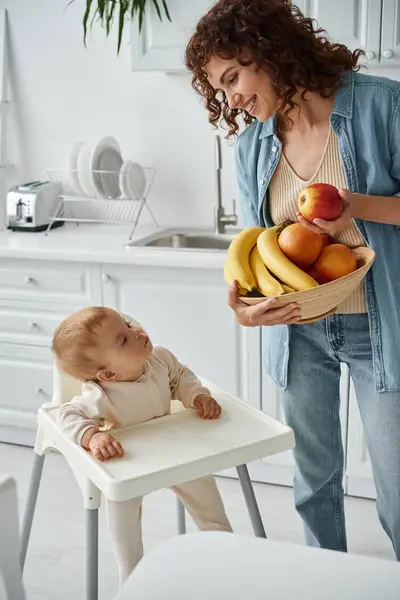 Criança bonita na cadeira do bebê olhando para a mãe com tigela de frutas frescas na cozinha, tempo de café da manhã — Fotografia de Stock