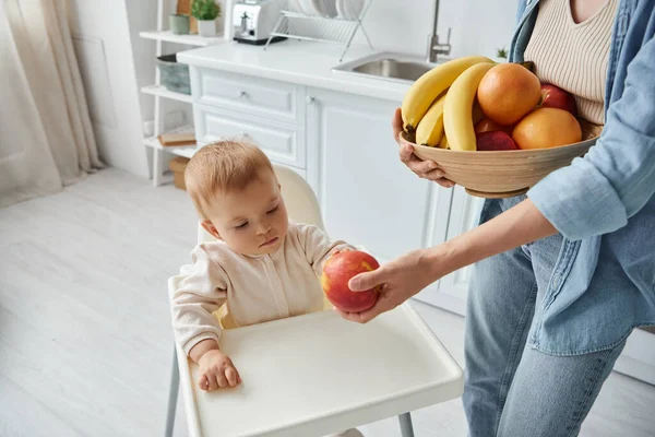 Mother with bowl of fresh fruits proposing ripe apple to little daughter sitting in baby chair — Stock Photo