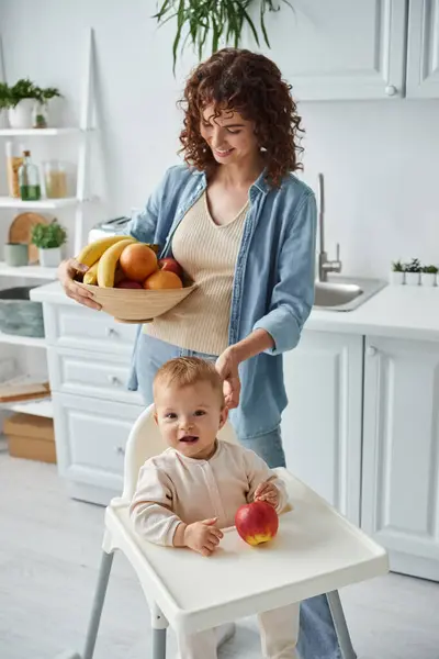 Niño emocionado sentado en silla de bebé cerca de manzana madura y mirando a la cámara cerca de mamá con frutas frescas - foto de stock