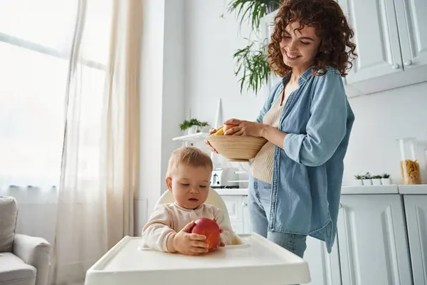 Mulher alegre com tigela de frutas frescas olhando para a filha da criança segurando maçã madura na cadeira do bebê — Fotografia de Stock
