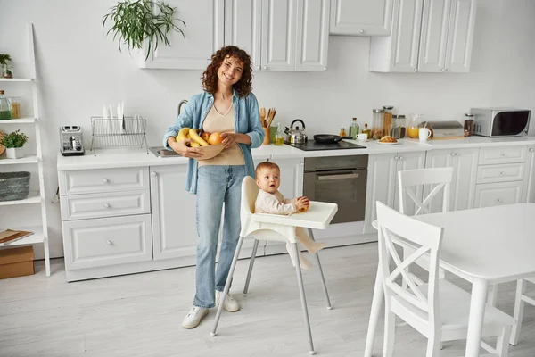 Pleased woman with fresh fruits looking at camera near toddler child in baby chair in modern kitchen — Stock Photo