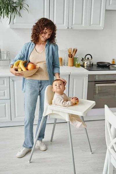 Smiling woman with fresh fruits stroking head of baby girl sitting in baby chair, morning in kitchen — Stock Photo