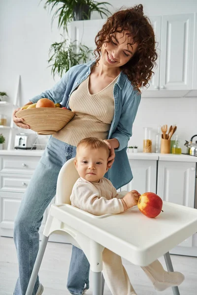 Joyful mother with fresh fruits stroking head of baby daughter sitting in baby chair near ripe apple — Stock Photo