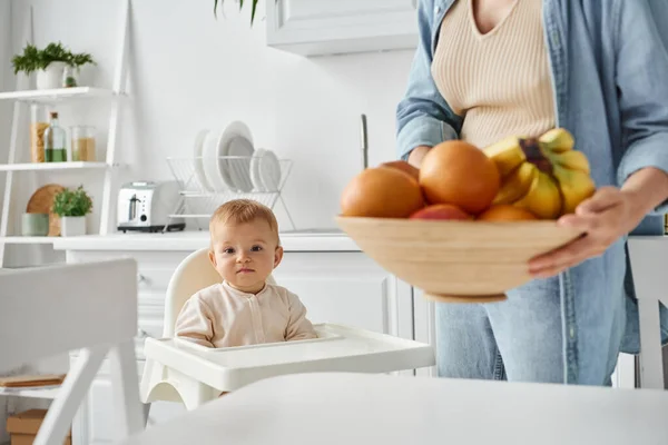 Little kid in baby chair looking at camera near mom with bowl of fresh fruits on blurred foreground — Stock Photo