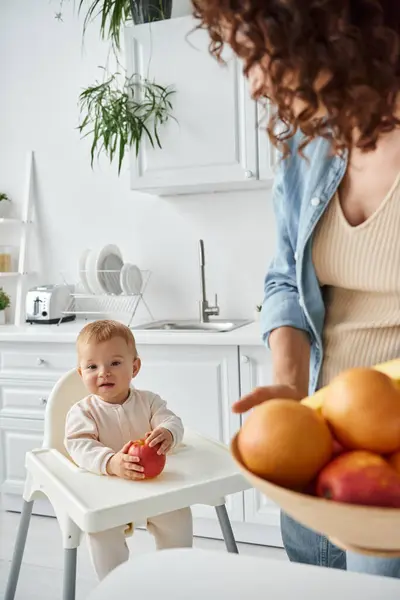 Joyful child sitting in baby chair with ripe apple near mom with fresh fruits on blurred foreground — Stock Photo