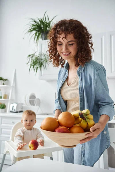 Glückliche Frau mit Schale mit reifen Früchten in der Nähe von Kleinkind Tochter sitzt im Babystuhl in der Morgenküche — Stockfoto