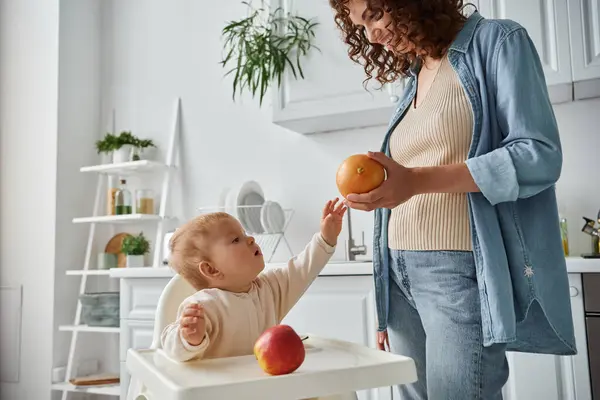 Милый ребенок в детском кресле достижения спелых оранжевый в руке улыбающейся матери на кухне, время обеда — стоковое фото