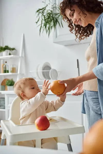Smiling mother giving fresh orange to little daughter in baby chair in kitchen, healthy breakfast — Stock Photo