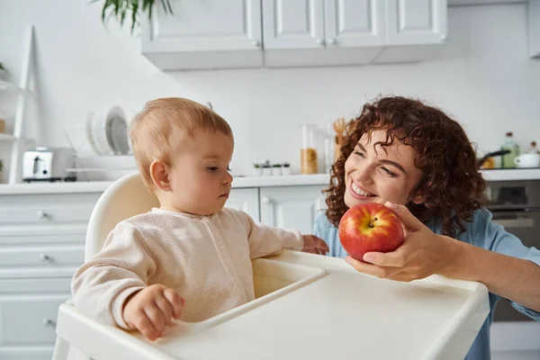 Cheerful woman proposing ripe fresh apple to adorable child sitting in baby chair in kitchen — Stock Photo
