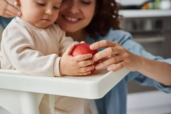 Little child holding ripe fresh apple while sitting in baby chair near happy mom, morning mealtime — Stock Photo