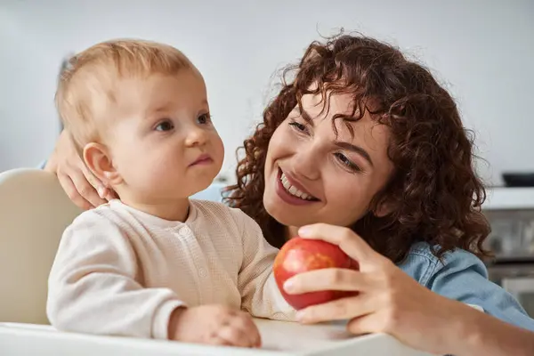 Delighted woman holding ripe apple near toddler daughter sitting in baby chair during breakfast — Stock Photo
