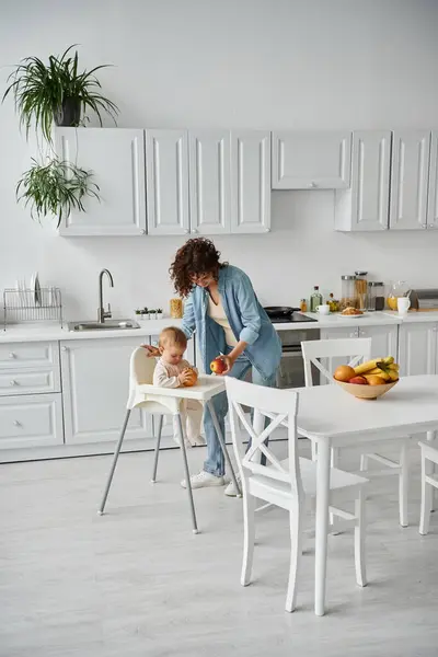 Mother and child in baby chair holding apple and orange near table with fruits in modern kitchen — Stock Photo