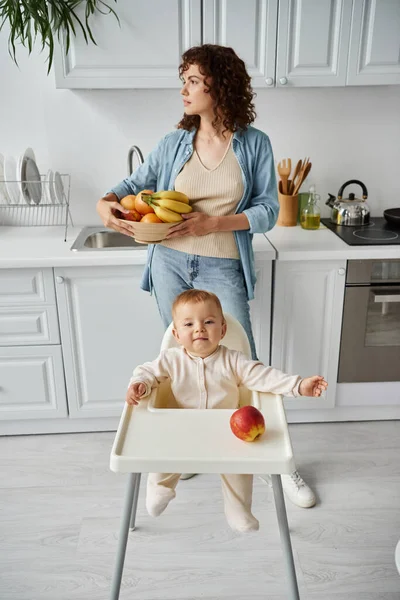 Mujer pensativa con frutas frescas mirando hacia otro lado cerca de niño juguetón sentado en silla de bebé cerca de manzana - foto de stock
