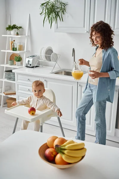 Happy woman with jug of fresh orange juice near kid in baby chair and ripe fruits in modern kitchen — Stock Photo