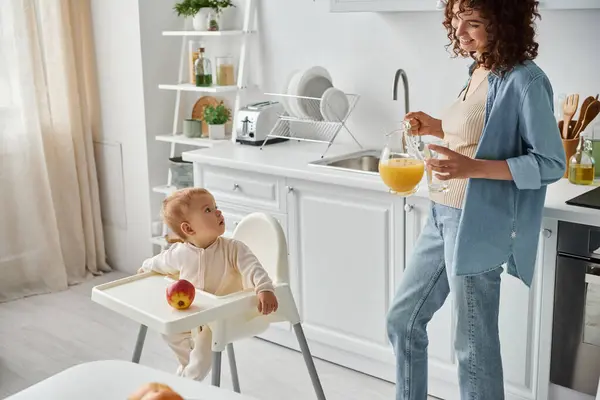 Niña pequeña sentada en silla de bebé cerca de manzana y mirando a mamá sonriente con jugo de naranja fresco - foto de stock