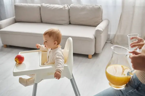 Маленький ребенок сидит в детском кресле рядом спелое яблоко и мать со свежим апельсиновым соком на кухне — стоковое фото