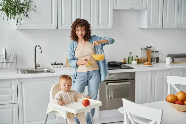 Fröhliche Frau gießt Orangensaft neben kleiner Tochter im Babystuhl mit reifem Apfel, Frühstück — Stockfoto