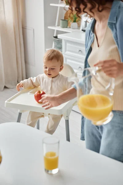 Woman with jug of orange juice pointing at ripe apple near little daughter sitting in baby chair — Stock Photo