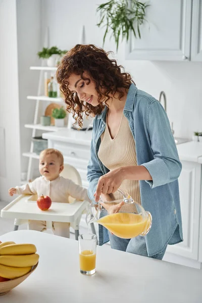 Smiling woman pouring fresh orange juice near child in baby chair in cozy kitchen, healthy breakfast — Stock Photo