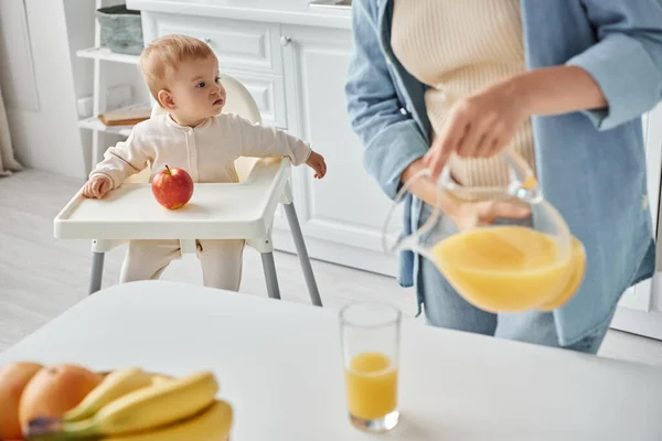 Femme versant du jus d'orange frais tandis que la petite fille assise dans la chaise de bébé près de pomme mûre — Photo de stock