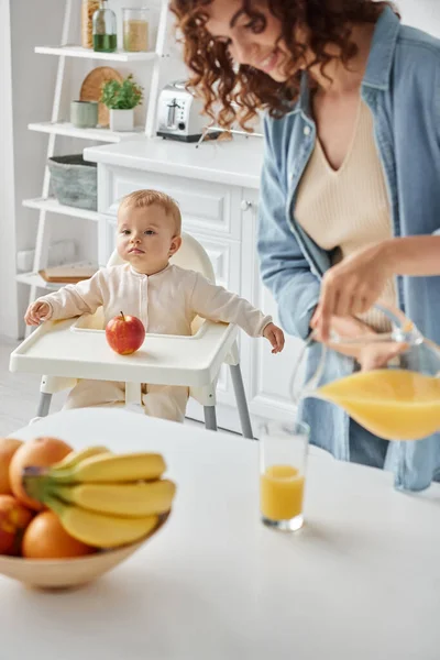 Cute child sitting in baby chair near apple while happy mom pouring fresh orange juice for breakfast — Stock Photo