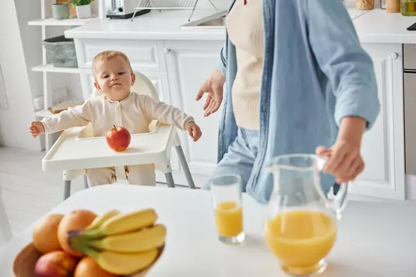 Cute kid in baby char looking at ripe apple near mother with jug of orange juice, morning in kitchen — Stock Photo