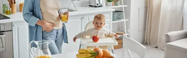 Menina na cadeira do bebê perto de maçã madura e mãe com copo de suco de laranja, tempo de café da manhã, banner — Fotografia de Stock