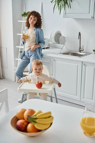 Joyful woman with glass of orange juice near toddler kid in baby chair and ripe fruits in kitchen — Stock Photo