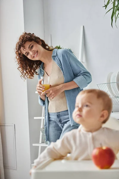 Улыбающаяся мать со стаканом апельсинового сока смотрит на дочку малыша в детском кресле рядом с спелым яблоком — стоковое фото