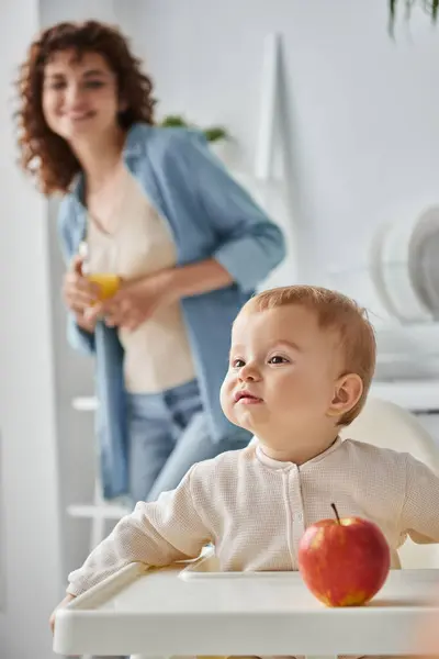 Bambina seduta vicino alla mela matura accanto alla madre con bicchiere di succo d'arancia su sfondo sfocato — Foto stock