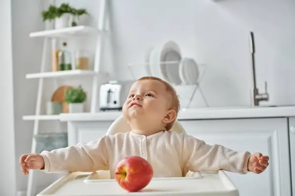 Tout-petit fille assise dans une chaise bébé près de pomme mûre et regardant loin dans la cuisine, enfance heureuse — Photo de stock