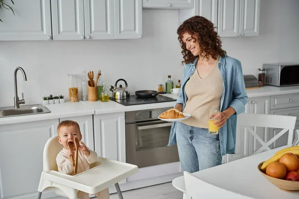 Smiling woman with croissant and orange juice looking at funny child chewing wooden tongs in kitchen — Stock Photo