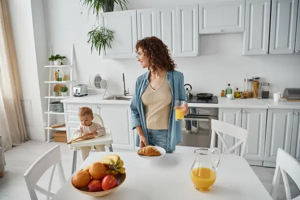 Woman with orange juice and croissant near fruits and child playing with wooden tongs in kitchen — Stock Photo