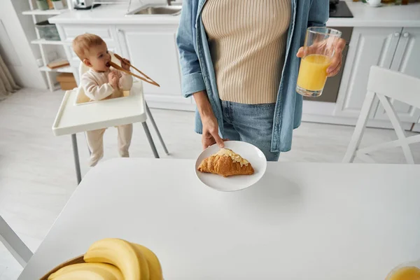 Woman holding fresh orange juice and croissant near toddler daughter playing with wooden tongs — Stock Photo