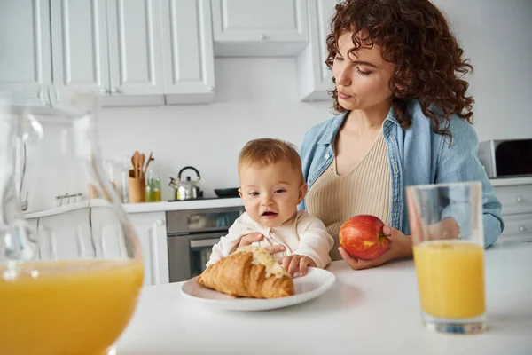 Mère et bébé près du croissant, pomme mûre et jus d'orange frais dans la cuisine, délicieux petit déjeuner — Photo de stock