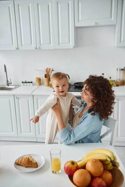 Femme tenant bébé gai près croissant et jus d'orange frais avec des fruits dans la cuisine, petit déjeuner — Photo de stock