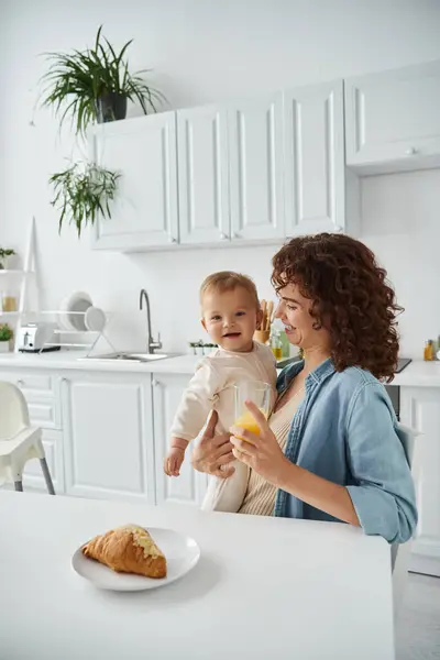 Mujer sonriente sosteniendo bebé feliz y vaso de jugo de naranja fresco durante el desayuno en la cocina moderna - foto de stock