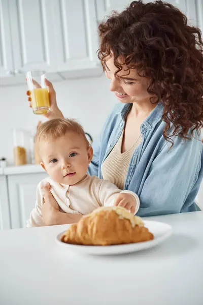 Cute baby looking at delicious croissant near mother with glass of orange juice, delicious breakfast — Stock Photo